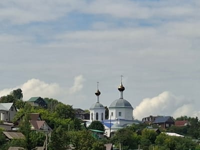 This media file captures a picturesque view of a village nestled on a hillside under a partly cloudy sky. The main subjects are two white churches with golden domes, standing prominently against the backdrop of lush green trees and scattered houses. The architecture of the churches, with their distinctive domes and crosses, suggests a religious or historical significance.
The scene is set in a rural environment, likely a village, given the presence of residential buildings with varied roof styles and the dense foliage. The houses appear to be of traditional construction, with some featuring colorful roofs, adding to the rustic charm of the location.
The activity or event taking place is not explicitly depicted; the image seems to be a still shot of a serene landscape. The time of day appears to be daytime, likely mid-morning or afternoon, given the brightness of the sky and the illumination of the buildings and vegetation. The weather is fair, with a mix of blue sky and white, fluffy clouds, suggesting a pleasant day.
Notable details include the vibrant green of the trees, which are in full leaf, indicating a season of growth, possibly late spring or summer. The hillside setting provides a natural elevation, offering a commanding view of the village and the surrounding landscape. The churches, with their white facades and golden domes, are a focal point, their spires reaching towards the sky. The scattered houses are interspersed with the trees, creating a harmonious blend of human habitation and nature. There is no visible text in the image. The overall mood conveyed by the image is one of tranquility and natural beauty.