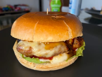 The image shows a close-up, eye-level shot of a cheeseburger on a dark plate. The burger is held together with a toothpick inserted through the top bun. The burger is thick, with a golden-brown bun, melted cheese, lettuce, ketchup, and what appears to be a meat patty. The background is blurred, showing a tabletop with a colorful object and some containers, suggesting an indoor setting. The lighting is somewhat dim and appears to be artificial. There is no visible text in the image. The focus is entirely on the burger, emphasizing its texture and ingredients.
