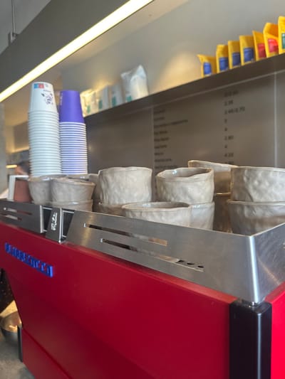 The image depicts the interior of Nomad Coffee Lab & Shop, located at Calle Hernán Cortés, 29, 39003 Santander, Cantabria, Spain. The central focus is on the upper part of a red commercial espresso machine, which has "NOMAD COFFEE" inscribed in blue letters on its front panel. A stainless steel rack with horizontal slots sits atop the machine, holding a collection of light beige, textured ceramic cups and bowls, some stacked and some arranged individually.
Behind the machine, a stainless steel back wall or counter area is visible. On the left, two stacks of disposable paper cups are present: one stack of white cups with a faint blue graphic, and another of purple cups with white horizontal stripes. Further back on a shelf, several white packaged items are neatly arranged. To the right of the paper cups, a menu is displayed on the stainless steel surface, listing coffee beverages and prices in Spanish, with entries like "CAFÉ", "AMERICANO", "ESPRESSO", and "LATTE" visible alongside numerical prices such as "2.30" and "2.80". Above this menu, a row of yellow, stand-up pouches with blue labels, likely containing coffee beans or products, are arranged on a higher shelf. The scene is illuminated by an overhead linear light fixture. No individuals are clearly visible, although a blurred head can be faintly discerned behind the paper cups in one cropped view, indicating a possible staff member or customer in the background.