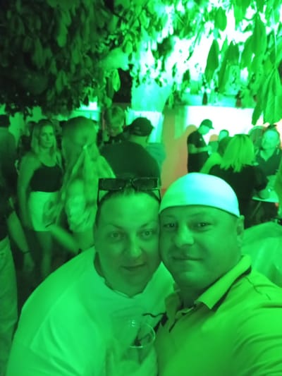 The image captures a group of people in an outdoor, dimly lit setting, illuminated by vibrant green light. In the foreground, two individuals are prominently featured. The person on the left, facing the camera, is wearing a white t-shirt and has dark hair. The person on the right, also facing the camera, is wearing a yellow polo shirt and a white head covering. Both individuals appear to be posing for a selfie. Behind them, several other people are visible, some in the background and others in mid-ground, engaged in what appears to be a social gathering. The environment suggests a nighttime event, possibly a party or festival, with foliage overhead casting shadows and contributing to the overall atmosphere. The location is identified as Hergiswil (NW), Switzerland.