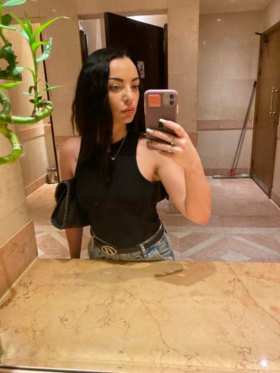 A woman with long, dark, wavy hair is taking a mirror selfie in what appears to be a hotel bathroom. She is wearing a black ribbed tank top, light-wash denim jeans, and a black belt with a gold buckle. A black purse with a chain strap is slung over her left shoulder. Her left hand, adorned with rings and painted nails, is holding a purple phone up to take the photo. Her expression is neutral. The bathroom features marble countertops and floors, with a large mirror reflecting the scene. A potted plant is visible on the left side of the image. In the background, a doorway and a mop are visible.
