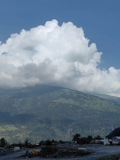 The image depicts a mountainous landscape near Solang, India. In the foreground, a dirt road is visible with scattered rocks, a yellow tarp, and a bicycle. A white concrete block is present on the left side of the road. The middle ground is dominated by a lush green mountainside covered with dense vegetation and trees, with some evidence of terraced farming. The background features layers of mountains fading into the distance, partially obscured by clouds. A large, prominent white cloud formation occupies the upper portion of the sky, with patches of blue sky visible. The overall scene suggests a natural, rural environment.