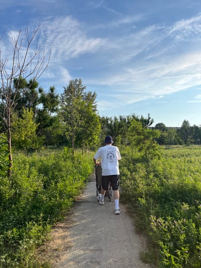 The image shows a person from behind, wearing a white t-shirt with text, dark shorts, white socks, and sneakers, pushing a stroller on a dirt path. The path is bordered by lush green vegetation and trees. The sky above is blue with scattered white clouds. The scene appears to be an outdoor recreational area. The location in the city of Kotovo, Russia is not discernible from the image alone.