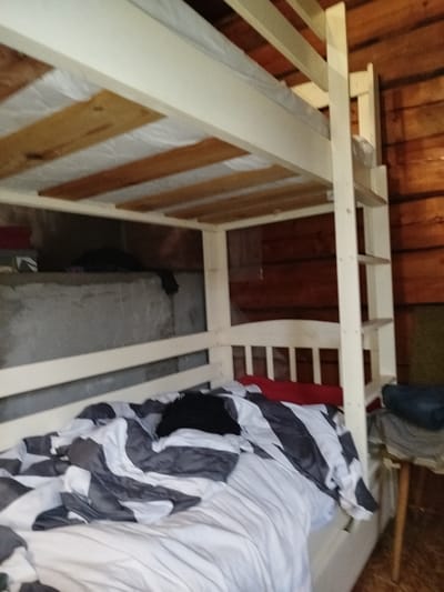 The image shows the interior of a room with a wooden-paneled wall and a bunk bed. The bunk bed frame is white, with wooden slats supporting the mattress. The lower bunk is partially visible and appears to have crumpled bedding with a black object on top. The upper bunk is also visible, with wooden slats and a mattress. The room has a rustic feel due to the wooden walls and the simple construction of the bunk bed. There is a concrete ledge visible under the upper bunk, with some items on it. The overall impression is that of a utilitarian living space, possibly in a rural or cabin-like setting within Дзешчанскі сельскі Савет, Belarus. There are no people visible in the image.
