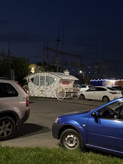 At night, a white ornate wagon, resembling a carriage, is parked on an asphalt surface. The wagon is decorated with gold scrollwork and has large spoked wheels. Above its entrance, a sign reads "COFFEE" with a crown emblem. Another red sign on the wagon says "ПРОДАЮ", indicating it is for sale. Several cars are parked around the wagon, including a silver SUV on the left and a white sedan behind the wagon. A blue car is in the foreground, partially obscuring the view of the wagon. In the background, industrial-looking power lines and structures are visible against the dark sky. The scene appears to be an outdoor area, possibly a parking lot or marketplace, in Vladikavkaz, Russia. No people are visible in the frame.