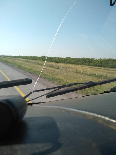 A view from inside a vehicle, looking out through the windshield on a clear day. The windshield has a thin white streak, possibly from a jet contrail, arcing across the upper portion of the sky. In the distance, a flat landscape with a line of trees borders a grassy field. A row of fence posts runs along the edge of the field next to a paved road with a double yellow line. The foreground shows the dashboard of the vehicle and the windshield wipers. The location is identified as Баймырза, Kazakhstan.