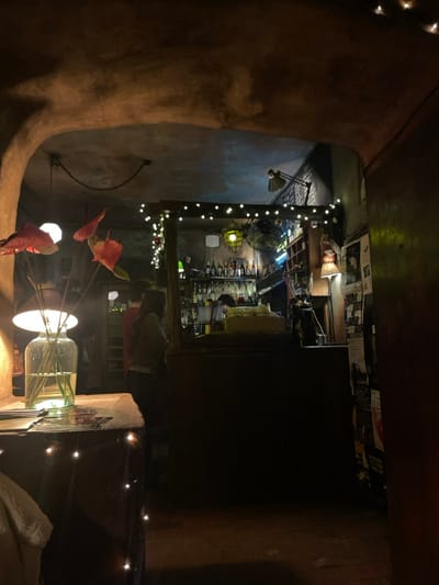 The image captures a dimly lit, cozy indoor space, likely a bar or cafe, at night. The scene is dominated by warm, artificial lighting, creating a relaxed and intimate atmosphere.
In the center-right, a bustling bar area is visible. A bartender, seen from the waist up, is actively working behind the counter, which is lined with numerous bottles of alcohol and bar equipment. Above the bar, several posters or flyers are affixed to the wall, though specific text is mostly illegible due to the distance and low light; fragmented words like "WIADOM" or similar characters can be vaguely discerned on some. The bar structure is adorned with strings of small, glowing white lights, and a unique, caged yellow-green light fixture hangs near the counter. A fan is also visible in this area.
To the far left, a large, organic-shaped, earthy-toned pillar or wall section dominates the foreground, adding to a rustic aesthetic. In the midground, a large glass jar or vase with reeds or sticks stands on a surface, illuminated from within by a light source. Several reddish, artificial leaf-like decorations, resembling anthuriums, are arranged around this vase. Another string of white lights is visible closer to the viewer on the right side. A person is partially visible seated at a table near these decorations, their back mostly to the camera. Further back, a table lamp with an adjustable arm casts light onto a nearby wall.
The overall ambiance is one of comfortable darkness, punctuated by numerous points of warm light from decorative string lights and various fixtures, suggesting a popular, atmospheric establishment, possibly a cellar bar given the rustic architecture hinted at by the large pillar. The presence of patrons and a working bartender indicates an active evening.