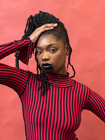 A woman with dark skin and braided hair poses against a solid salmon backdrop. She wears a form-fitting long-sleeved shirt with horizontal red and black stripes. Her makeup includes dark eyeliner and glossy black lipstick. Her left hand is raised to her head in a thoughtful pose, and she gazes directly at the viewer with a slight tilt of her head. The photograph is oriented at an angle, and the lighting is soft and even.
