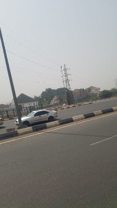 A silver car parks alongside a road with black and white curbing. Power lines run parallel to the road, extending to the left. A few buildings stand among trees in the background, with a larger structure visible further in the distance. The asphalt road stretches across the right portion of the image, displaying a faint dividing line. The sky is bright and hazy.