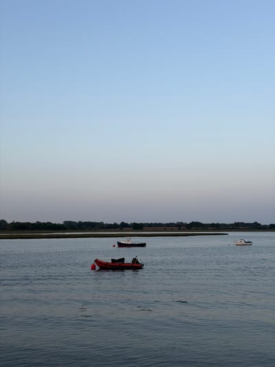 The image depicts a waterway in East Suffolk, United Kingdom, with a sliver of land running vertically through the middle of the frame. The left side of the image shows a sky gradient from light blue at the top to a light grey near the land. The right side shows water with small waves. Three boats are visible on the water: a larger, light-colored boat towards the top, a smaller white and red boat near the middle, and an orange and black boat near the bottom. The land is lined with trees.