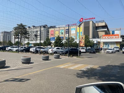 The image depicts a street scene in Andijan City, Uzbekistan, outside a shopping center. In the foreground, a paved road with a yellow line is visible, leading towards a pedestrian crossing with yellow and white stripes. Two large, circular concrete planters are positioned on the side of the road. Beyond the planters is a parking lot filled with various cars, including white sedans, black sedans, and a white SUV. The background features a multi-story shopping center with several prominent signs. One sign reads "korzinka" in red letters, another "HONOR," and a large orange sign displays "indenim." Several other advertisements and signs are visible on the facade of the building, some featuring images and text in what appears to be Uzbek. A few trees are scattered around the scene. Overhead, strings of small lights are suspended across the sky. The sky is clear and blue. In the far background, parts of other buildings and a construction crane are visible. No people are clearly discernible in the image.