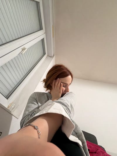 This low-angle indoor photograph, taken in Reims, France, features a young person with reddish-brown hair. The subject is positioned above the camera, leaning or looking downwards, with one hand partially covering their face, obscuring their full expression. They are dressed in a grey hooded sweatshirt, with a white layer visible underneath, and dark leggings or pants. A silver bracelet adorned with star and heart-shaped charms is clearly visible on their left wrist/forearm, which is closest to the camera due to the perspective.
The background consists of plain white walls and ceiling. Below the subject, a modern white window frame with two sections, each featuring closed horizontal blinds, is visible. On the narrow window sill above the glass, a thin yellow object (possibly a pencil or straw) and a small, clear glass or plastic item can be seen. In the upper right corner, part of a dark furniture piece, possibly a couch or chair, is visible with a reddish textile draped over it. The room is artificially lit, providing no indication of the time of day or external weather conditions. No text is visible within the image. The unique perspective creates a sense of the subject looking down at the viewer or camera.