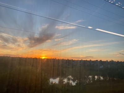 This image depicts a serene landscape at sunset, viewed from a window, likely from a moving vehicle given the streaks and reflections. The sky is a vibrant canvas of blues and oranges, with wispy clouds catching the warm light of the setting sun. A prominent feature in the sky are the distinct lines of overhead wires, suggesting an urban or semi-urban environment.
Below the sky, a dense line of trees forms a dark silhouette against the horizon, with a small body of water reflecting the sky's colors. Beyond the trees, a town is visible, with buildings and structures dotting the landscape. The warm glow of the sunset illuminates the scene, casting a peaceful and tranquil mood. The specific time of day is clearly sunset, evidenced by the color of the sky and the low angle of the sun. The weather appears clear, with minimal cloud cover except for the wispy formations. There is no visible text in the image. The overall impression is one of natural beauty and quietude, possibly experienced during a journey.