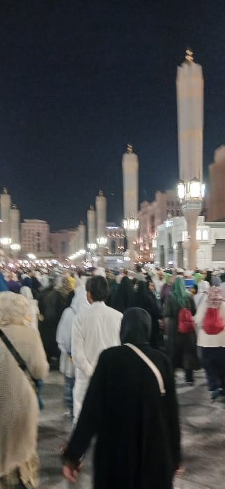The image captures a crowded outdoor scene at night in the city of Medina, Saudi Arabia, specifically in the plaza surrounding Al-Masjid an-Nabawi (the Prophet's Mosque). Multiple tall, white, cylindrical minarets, each topped with a golden finial, rise prominently against the dark sky. The area is brightly illuminated by numerous multi-lantern streetlights and lights emanating from the surrounding mosque buildings and structures. A dense congregation of people, primarily adults, is observed walking, mostly with their backs to the viewer, indicating movement through the space. Individuals are dressed in various forms of Islamic attire; men are seen in white thobes, and women wear head coverings, hijabs, and abayas in colors including black, beige, and green, with some carrying red backpacks. The ground appears to be a paved surface.