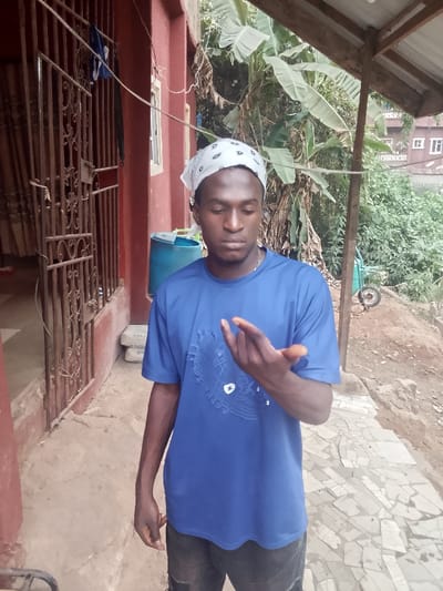 A dark-skinned man wearing a blue t-shirt and a white paisley bandana stands outdoors. He is looking down at his outstretched left hand. Behind him is a red building with a metal gate. A blue barrel is visible in the background. Lush green plants and a roof structure are also in the background. The ground is a patchwork of light-colored stone. The man's expression is serious and thoughtful.