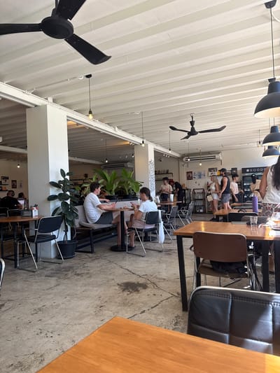 The image shows the interior of a cafe in Choeng Mon, Thailand. The cafe has a high ceiling with white exposed beams and industrial-style pendant lighting. Multiple black ceiling fans are visible. The flooring is concrete with visible cracks and stains.
In the foreground, a wooden tabletop and a black leather chair are prominent. Further into the cafe, several tables and chairs are occupied by patrons. On the left, a large potted plant stands near a white pillar.
People are seen sitting at tables, talking, and interacting. Some are looking at their phones. On the right side of the cafe, a counter area is visible with shelves stocked with goods. Several staff members appear to be working behind the counter. The overall atmosphere suggests a casual dining or coffee shop environment.