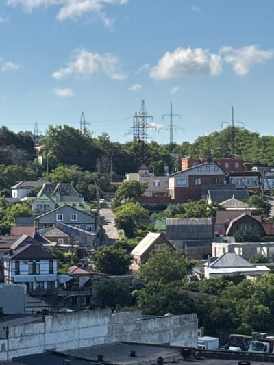 The image depicts a residential area on a hillside in Novorossiysk, Russia, with numerous houses and trees visible. In the background, high-voltage power lines with support towers stretch across the landscape. The houses are of varying sizes and architectural styles, many with pitched roofs. A paved road winds through the settlement, with a white car visible on it. The sky is blue with scattered white clouds. The scene appears to be a typical urban or semi-urban environment in a hilly region. There are no people visible in the image.