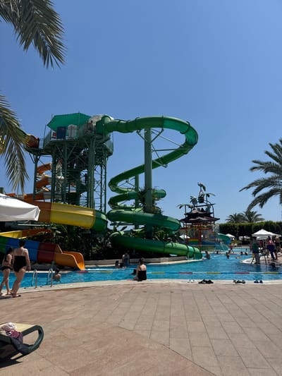 In Aksu, Turkey, a vibrant water park is captured on a sunny day. Dominating the scene are several large, colorful water slides. A towering green spiral slide curves downwards, juxtaposed with a yellow and orange slide descending from a multi-tiered structure. A smaller red and orange slide is also visible. The water park features a clear blue swimming pool where several people are seen relaxing and swimming. Along the pool's edge, lounge chairs and umbrellas are scattered. Palm trees frame the scene, adding to the tropical ambiance. People, dressed in swimwear, are scattered throughout the park, some entering the pool, others walking nearby. The environment is bright and cheerful, indicative of a recreational area.
