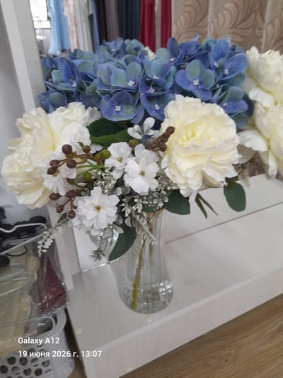 This is a close-up, eye-level shot of a floral arrangement in a glass vase. The arrangement features large, light yellow peonies and clusters of blue hydrangeas, interspersed with smaller white flowers and brown berries on thin stems. Green leaves are also visible, along with sprigs of delicate white foliage. The vase has a rippled texture and is filled with clear liquid. The flowers are placed on a white, reflective surface that appears to be a shelf or tabletop, with a wooden floor visible in the background. To the left of the vase, a white plastic basket containing plastic-wrapped items and what appear to be clothing hangers is partially visible. The background also shows a reflection of curtains and what looks like a window with blue drapes. A watermark at the bottom left corner reads "Galaxy A12 19 июня 2026 г. 13:07". The overall scene depicts a still life composition of artificial flowers.