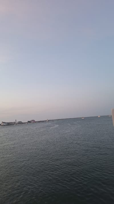 The image depicts a wide view of the ocean off the coast of Nantucket, Massachusetts, United States of America. The sky is a pale, cloudless blue, suggesting either early morning or late evening. The water is a deep, muted blue-green, with small ripples indicating a gentle breeze. In the distance, along the horizon, a line of buildings is visible, resembling a waterfront town or harbor. Several sailboats are moored or cruising on the water, varying in size and distance. A white lighthouse stands out among the buildings on the left side of the frame. The overall scene is tranquil and scenic, capturing the essence of a coastal environment.