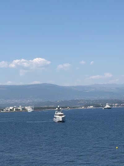 A wide-angle, eye-level, horizontal shot captures a serene coastal scene under a clear blue sky with scattered white clouds. The foreground is dominated by the deep blue, slightly rippled surface of the sea. In the middle ground, several white yachts are visible. One prominent yacht, a large, modern vessel with multiple decks and a sleek design, is positioned slightly right of center. Further to the left, a cluster of buildings, appearing to be hotels or apartment complexes, lines the shore, with a smaller yacht anchored nearby. Beyond the coastline, rolling hills recede into the distance, topped by a hazy mountain range. The overall impression is one of calm and luxury in a Mediterranean setting. This scene is indicative of the view from the sea towards the city of Théoule-sur-Mer, France, known for its beautiful coastline and upscale waterfront properties.