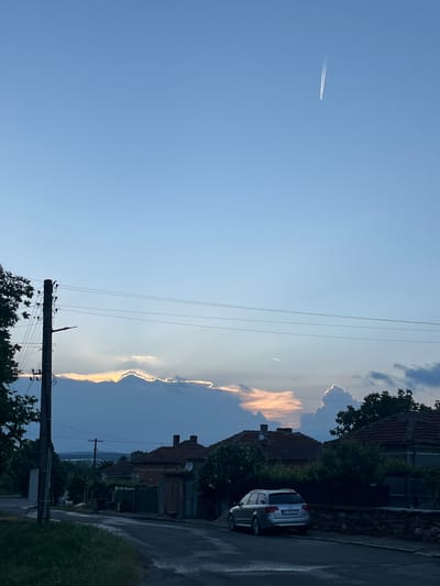 The image captures a street scene in Lozarevo, Bulgaria under a blue sky. A contrail from an airplane is visible in the upper left quadrant. Overhead power lines run across the top half of the frame, connecting to a utility pole in the lower center. To the right of the utility pole are houses with red tiled roofs, interspersed with greenery. A silver-colored car is parked on the right side of the street. Golden light illuminates the edges of the clouds adjacent to the houses, suggesting it is near sunset. The street surface appears wet.