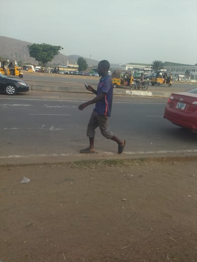 The image shows a man walking on a sidewalk along a busy road in Maraba, Nigeria, at coordinates 9.024 latitude and 7.573 longitude. The man is wearing a blue t-shirt, khaki pants, and sandals. He is walking with his left foot forward, and his right arm is extended as he holds a smartphone in his hand. The man is looking down at the phone, and his expression is neutral. The road is asphalt and has a white dividing line. The sidewalk is dirt and gravel. The man is walking towards the left side of the frame. In the background, there are numerous cars, motorcycles, and buildings. On the opposite side of the road, a red car can be seen driving towards the camera. The man's action suggests he is either making a call or checking a message on his phone. The scene is typical of a bustling urban area, with people and vehicles constantly on the move.