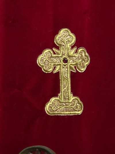 On a red surface, a gold Orthodox cross with a red gem at the center is positioned horizontally. Intricate scrollwork embellishes the cross. The location of Balsha, Bulgaria is not inferable from the image.