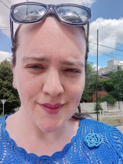 A woman, likely of European descent with light skin and dark hair pulled back, is captured in a close-up, selfie-style portrait. She is wearing dark-rimmed sunglasses, and her blue eyes are visible through them. Her lips are painted with a matte pink lipstick. She is wearing a bright blue, intricately crocheted top with a scalloped neckline and a blue crocheted rose brooch on her left shoulder. The background reveals a bright, cloudy sky with some sunlight glinting off the sunglasses. Behind her, there are trees, a fence made of white brick, and a building with a red roof and white structures. The environment appears to be outdoors in a city setting. Based on available visual information, the setting is the city of Kherson, Ukraine.