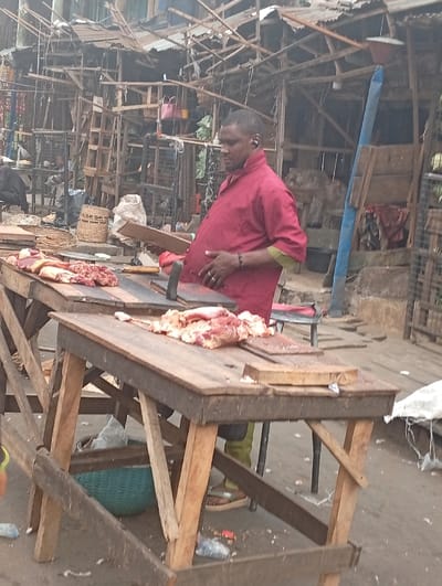 This image captures a vibrant scene from an outdoor market, likely specializing in meat. In the foreground, an adult man, possibly a butcher or vendor, stands behind two sturdy, rustic wooden tables. The man has a dark complexion, short dark hair, and is wearing a maroon-red short-sleeved shirt with light green cuffs. He appears to be wearing light green boots or shoes with sandals and has an earbud in his right ear. A ring is visible on his left ring finger, and his hands are resting over his midsection. His expression is neutral, looking downwards or slightly to his left.

The two wooden tables in front of him serve as display surfaces for raw red meat. Various cuts of meat, some with bone, are laid out on both tables. On the left table, a large cleaver or knife is positioned next to the meat. The right table features a wooden chopping block. Beneath the left table, a light green woven basket is visible. The ground around the tables is unpaved, appearing to be dirt or rough concrete, and is scattered with litter including plastic bottles, wrappers, and what appear to be small scraps or bone fragments.

The background of the image reveals a dense cluster of makeshift market stalls and structures. These are constructed from rough wooden planks, beams, and corrugated metal sheets, giving them a somewhat aged and informal appearance. Numerous wooden supports and frames are visible, creating a complex, multi-layered backdrop. Among the background elements, a pink bag hangs from a structure on the left, and a tall blue pole or beam stands prominently on the right side of the frame. The overall impression is that of a bustling, traditional marketplace.

The lighting suggests it is daytime, possibly overcast or during the mid-morning/afternoon, as there are no harsh shadows. The market appears to be a busy commercial hub. No discernible text is visible within the image.