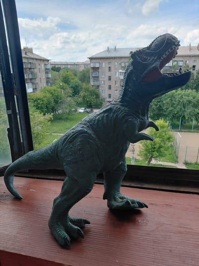 A dark green, toy Tyrannosaurus Rex figure stands on a reddish-brown wooden surface, positioned in front of a window overlooking a city. The dinosaur toy is in profile, facing right, with its mouth open and head tilted upward. The background visible through the window shows multi-story apartment buildings, lush green trees, and a cloudy sky. A sports field with a fence can be seen in the distance. The scene is likely located in Magnitogorsk, Russia, given the architectural style of the buildings. The lighting suggests it is daytime, possibly late morning or early afternoon.
