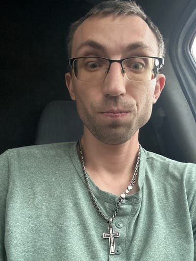 A man in his 30s, wearing a pale green henley shirt and glasses, is photographed from the chest up. He is looking directly at the camera with a neutral expression. A silver chain with a cross pendant rests on his shirt. The background is dark, suggesting an interior space, possibly a vehicle, with a glimpse of a grey seat on the left. There is no discernible location within the city of Berezivka, Ukraine.