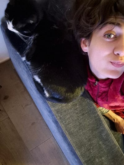 A person reclines on a gray denim-upholstered surface with a black and white cat nestled on their shoulder. The person wears a red garment. The cat's head is visible, showing its black fur and white markings on its face and chest. The foreground shows a wood floor. The person looks up at the camera.