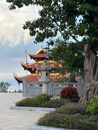 The image depicts a multi-tiered Buddhist pagoda with red tiled roofs and golden accents, situated in the city of Diên Khánh, Vietnam. The pagoda is partially obscured by lush green foliage from a large tree with exposed roots in the foreground, and other trees and bushes. Prayer flags in various colors are visible, adorning the pagoda and surrounding structures. A stone lantern stands prominently in front of a decorative stone wall. The ground is paved with light-colored square tiles. The sky is partly cloudy.