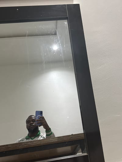 A person is reflected in a large, framed mirror with a black frame. The person is holding a blue smartphone and appears to be taking a photograph of themself. The person is wearing a white and green striped jacket. The background is a plain white wall. The location is unspecified, and there is no indication of it being in Iddo Tudunwada, Nigeria.