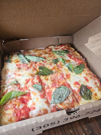 The image depicts a slice of pizza in a cardboard box. The pizza has a tomato sauce base, topped with cheese and fresh basil leaves. The box has a phone number "(305) 396-724" printed on the side. Key Largo is a part of the (305) telephone area code, so that would be the location where the pizza was bought.
