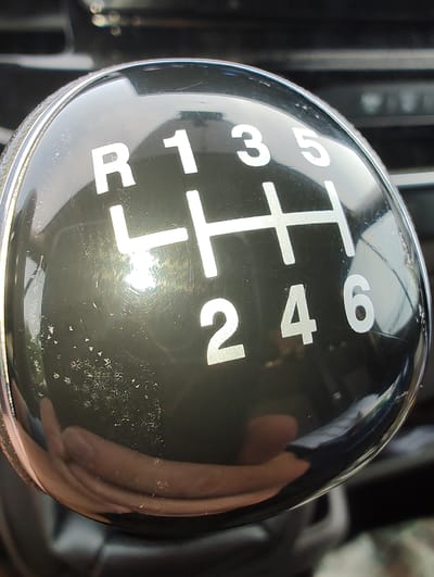 A close-up, high-angle shot shows the glossy black shift knob of a manual transmission vehicle, reflecting a hand holding it. The shift knob has white markings indicating the gear pattern: R, 1, 3, 5 on the top row, and 2, 4, 6 on the bottom row. The background is blurred and dark, suggesting an indoor environment such as the interior of a car. The image is focused on the shift knob, with a slight bokeh effect in the background. There is no indication of the location being in Wangels, Germany.