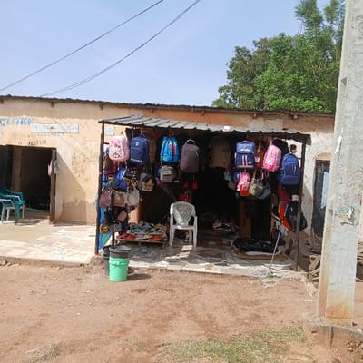 The image shows a small storefront in Kufang, Nigeria, located at coordinates 9.863 latitude and 8.867 longitude. The store is a simple structure with a corrugated metal roof and a dirt floor. It is open to the front and has a variety of bags hanging from a metal rack. There are also several bags and purses stacked on a wooden table inside the store. There is a plastic chair in front of the store and a green bucket to the left. The store is located on a dirt road. The surrounding area is undeveloped and appears to be a residential neighborhood. The sky is clear and blue, with a few wispy clouds. The image suggests that the store is selling a variety of bags and purses for everyday use.