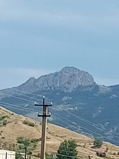 The image shows a wide shot of a mountainous landscape under a clear blue sky. In the foreground, a utility pole with crossbars and insulators stands to the right of center, with power lines extending horizontally across the frame. The pole is made of wood. The terrain in the mid-ground consists of a dry, rolling hillside with sparse vegetation, appearing arid and possibly scrubland. In the background, a large, rugged mountain range dominates the scene, with a distinct, rocky peak. The slopes of the mountain range are covered in dark green vegetation. The overall impression is of a rural or natural environment. The specific location of Щебетовский поселковый совет, Ukraine, is not discernible from the visual content of the image itself.