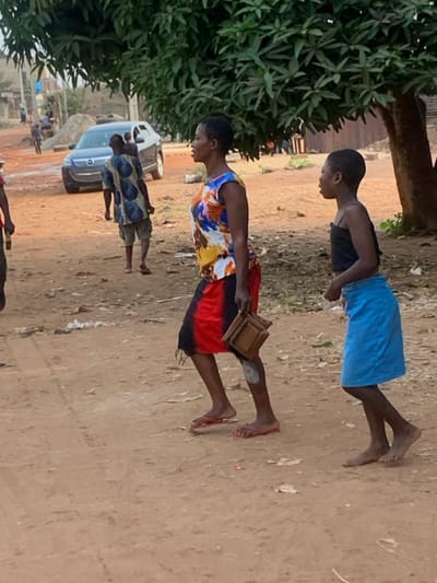 A scene captures three individuals, seemingly in an outdoor setting. A woman in a bright, multicolored top and red skirt carries a wooden tray. Beside her, a girl wears a black top and blue skirt. Another person is visible behind the woman. The setting appears to be a dirt road in a rural area with sparse vegetation, a car, and other figures in the distance. The people are walking barefoot.