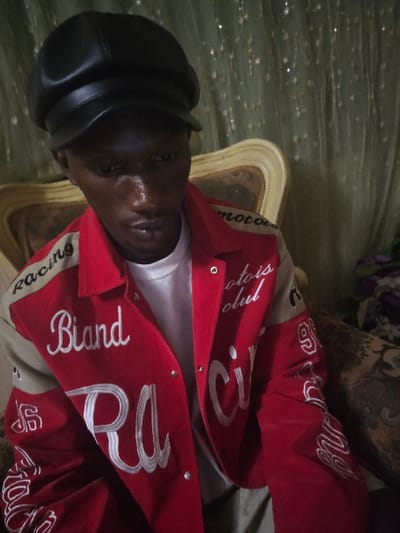 A person wearing a black hat and red racing jacket reclines in a chair. The jacket features white embroidered script, including "Biand," "Racing," "Motors club," and numbers like "13" and "95." The chair has a light-colored frame. The background includes a textured surface, possibly a wall or fabric. The image appears to be indoors with a slightly muted color palette.