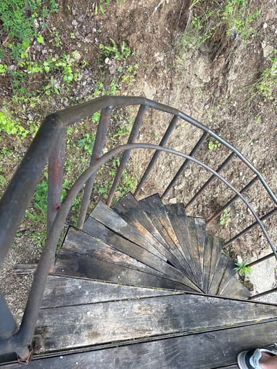 An outdoor, top-down shot shows a rustic, spiral staircase constructed of weathered wooden planks and a dark metal railing. The staircase is set against a backdrop of dry, rocky earth and sparse green vegetation. The wooden treads are worn and uneven, with some appearing damp and mossy. The metal railing is also aged, showing signs of rust. The surrounding ground is covered in dirt, small stones, and patches of grass and small weeds. In the lower right corner, the edge of a person's foot wearing a New Balance sneaker is visible on one of the wooden planks, indicating someone is standing on or near the staircase. The scene evokes a sense of disuse and natural integration, suggesting the staircase may be part of an older structure or an access point to a natural area. There is no specific information provided within the image to pinpoint its exact location within the city of Lyady, Russia.