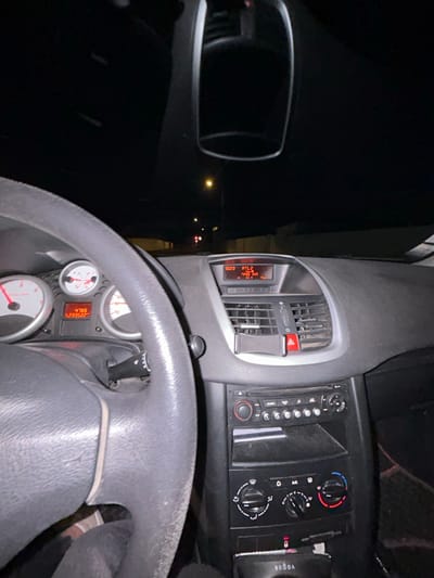 An interior car view features the steering wheel, dashboard, and side mirror. The dashboard includes air vents, a digital display showing "12:23 RTL2 44.0°C", and climate control buttons. A phone holder is mounted on the vents. The steering wheel houses instrument gauges, including a speedometer. The side mirror reflects a dark exterior environment with faint lights, possibly on a road at night. A "Vogue" branded object is located on the right.