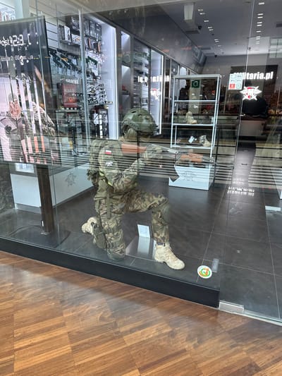 The image shows the storefront of a military surplus store, likely located in Łódź, Poland, given the context provided. The store's window display features a mannequin dressed in camouflage military attire, kneeling and holding a rifle. The mannequin is equipped with a helmet, tactical vest, and a holster on its leg. Behind the mannequin, shelves are visible displaying various military-style items, including what appear to be replica firearms, sunglasses, and other accessories. The store's signage, visible in the reflection on the glass, includes "militaria.pl," a website for a Polish military surplus retailer. The flooring inside the store appears to be dark tile, while the flooring outside is a polished wooden parquet. The scene is well-lit, suggesting it is daytime.