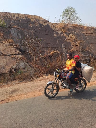 In Itobe, Nigeria, a daylight scene depicts a motorcycle with two occupants and cargo on an asphalt road. A male driver, wearing a yellow and red patterned short-sleeve shirt, plaid shorts, and blue sandals, operates a silver-fendered motorcycle. A female passenger, dressed in a black top, black pants, a red headwrap, and white flip-flops, rides pillion. A large, light-colored sack is secured to the rear of the motorcycle. To the left, a dirt shoulder abuts the road. In the background, a large, weathered, dark brown and tan rocky embankment rises sharply, showing horizontal striations and sparse, dry vegetation. A single, small green-leafed tree stands atop the embankment against a clear, light blue sky.