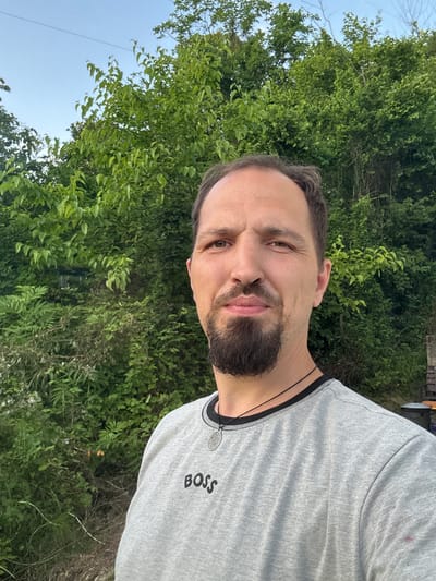 The image is a selfie of a man in his late 30s to early 40s, with dark hair, a beard, and a goatee. He is wearing a grey t-shirt with the word "BOSS" printed on it in black lettering, and a silver pendant necklace. He is standing in front of a dense wall of green trees and foliage, with a clear sky above. The location is identified as Verkhniy Yurt, Russia. The overall lighting suggests it is daytime, possibly late afternoon or early evening, given the soft light and absence of harsh shadows. The background indicates an outdoor, natural setting, likely a rural or suburban area. There are no other individuals or significant objects in the immediate vicinity.