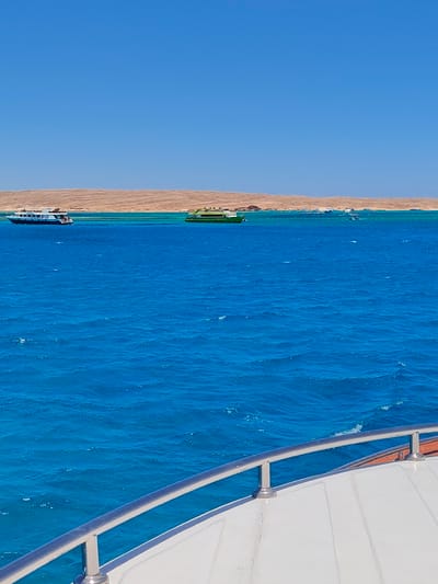 This media file depicts a bright, sunny day at sea, likely in Hurghada, Egypt. The primary subjects are several boats on the intensely blue, rippling water. In the foreground, the railing and deck of a white boat are visible, suggesting the viewer is on a vessel.

In the mid-ground, a long, white and blue passenger boat is sailing, with several people visible on its deck. Further to the right, a vibrant green boat with a similar passenger capacity also navigates the water. In the distance, a line of various smaller boats is present, some appearing to be moored near a sandy, arid coastline. The coastline itself is characterized by low, undulating hills with a tan, desert-like appearance, indicative of an Egyptian coastal landscape.

The sky is a clear, vibrant blue, with no clouds, suggesting fair weather. The sunlight is strong, casting a bright illumination on the scene. The water's color transitions from a deep, rich blue to a lighter, turquoise hue closer to the shore and around the coral reefs, hinting at shallow, clear waters. The overall mood is serene and recreational, suggesting a typical tourist excursion or a peaceful day out on the Red Sea. No visible text is present in the image.