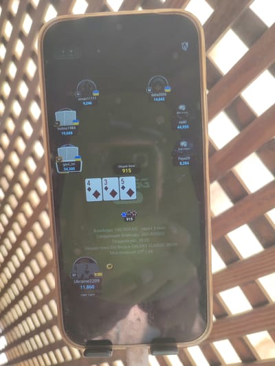 The image shows a close-up of a smartphone screen displaying a poker game interface, likely an online application. The phone is placed on a surface, and the background suggests an outdoor setting with a slatted wooden structure. The phone screen displays player avatars, usernames, scores, and card suits. Specific player names visible include "viman11111", "holms1984", "daha2026", "naiki", "gavi_nn", "Feya29", and "Ukraine2209". The game appears to be in progress with cards displayed on the screen, and information about blinds and pot size is also visible. There are no people clearly visible in the frame, other than the digital representations on the screen. The overall environment appears to be a casual outdoor setting. There is no information within the image that can be used to determine its specific location within Santa Lucía de Tirajana, Spain.