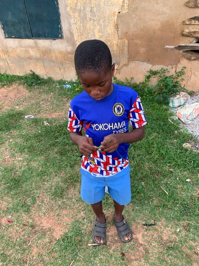 A young Black boy stands on a patch of grass and dirt outside a dilapidated building. He is wearing a blue Chelsea FC jersey with the Yokohama Tyres logo and blue shorts. His hands are busy manipulating a green string. His feet are clad in dark grey sandals. The building behind him is made of weathered tan plaster with cracks and peeling paint. A dark green, diamond-patterned metal sheet is affixed to the wall to the left, resembling a small window or vent. To the right, the wall shows a hole with some wooden debris protruding. Several bags, possibly cement or fertilizer, are piled on the ground near the building. The ground is a mix of sparse green grass, dry brown patches, and loose dirt. Scattered pieces of white and colored debris are visible on the ground.