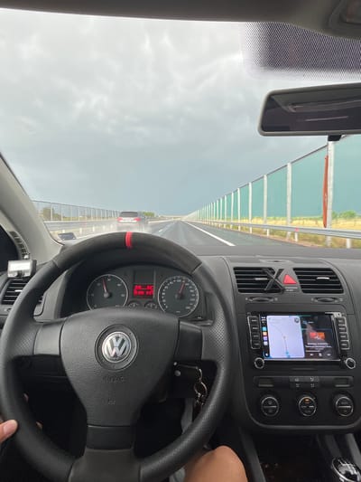 An internal view from a Volkswagen car shows the driver's perspective on a highway near Chokoba, Bulgaria. The image, taken during the mid-afternoon with an overcast sky, captures the dashboard, steering wheel, infotainment system, and the road ahead through the windshield.

In the foreground, a black, perforated leather steering wheel with a red accent stripe is visible, featuring the Volkswagen logo and "AIRBAG" text. The driver's right hand or arm is partially visible on the bottom right, suggesting the car is being driven. Behind the steering wheel, the instrument cluster shows a speedometer indicating a speed between 120 and 130 km/h, a tachometer, and a digital display with "15:32" (time), "16.5" (likely temperature in Celsius), "20", "1390", and "5427" (possibly trip or total mileage).

The center console features an infotainment system displaying a navigation map alongside music playback details, "Беатрис X MONKEY - Тинтири Минтири / Beat....". Buttons around the screen include "MENU", "PHONE", "NAV", "RADIO", "MEDIA", "MUTE", and "TONE". Below these are climate controls with "OFF", "DUAL", "AUTO", and "AC" buttons, and an "PASSENGER AIR BAG" indicator. A set of car keys hangs from the ignition or a nearby hook.

Outside the car, the highway extends into the distance under a grey, cloudy sky. The road appears damp or recently wet. A white sedan is visible some distance ahead. To the left, a sound barrier or guardrail with translucent green panels runs along the side of the highway, consistent with infrastructure found on European motorways. A rearview mirror is also visible at the top of the windshield. The overall scene depicts a neutral travel moment on a highway.